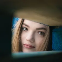 Portret młodej kobiety z bliska, spoglądającej przez niebieską drewnianą ramę, z delikatnym uśmiechem i jasnymi włosami.