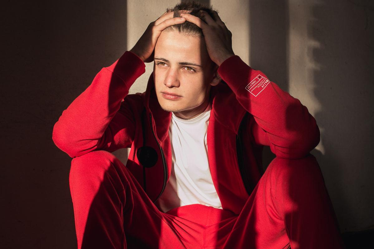 Młody mężczyzna w czerwonym dresie siedzi na podłodze, opierając dłonie na głowie, zamyślony wyraz twarzy, oświetlony promieniami słońca padającymi przez okno.