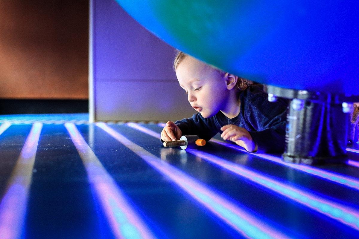 Mały chłopiec w ciemnym pomieszczeniu bawi się małą zabawką na podświetlanej, niebieskiej powierzchni z wyraźnymi, poziomymi liniami, z bliska, pod niskim kątem.