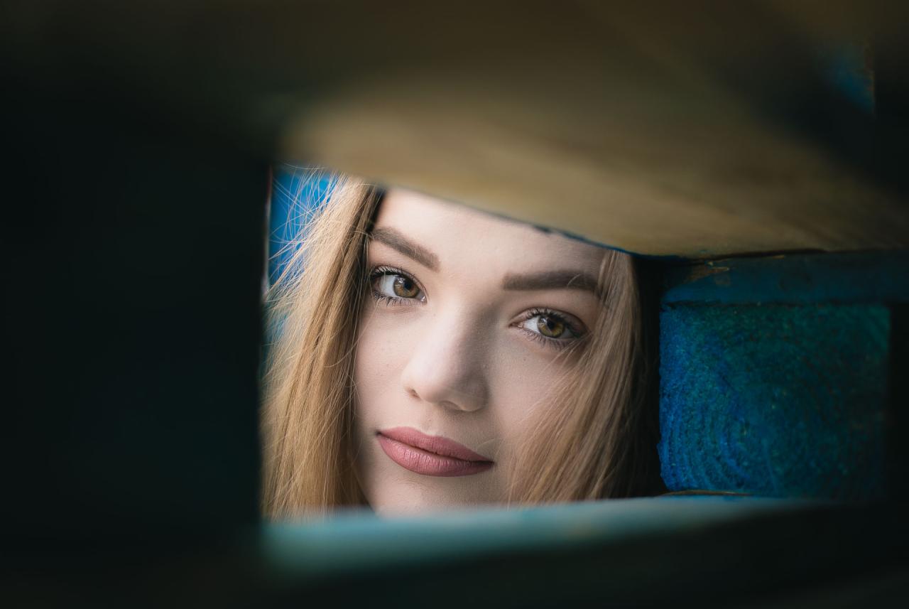 Portret młodej kobiety z bliska, spoglądającej przez niebieską drewnianą ramę, z delikatnym uśmiechem i jasnymi włosami.