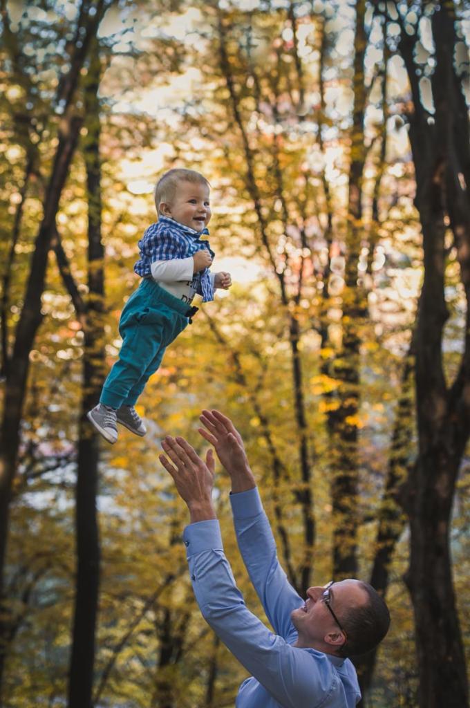 Uśmiechnięty niemowlak w niebieskiej koszuli w kratę i turkusowych spodniach unosi się w powietrzu, rzucony przez ojca w parku jesienią, z drzewami o żółtych liściach w tle.