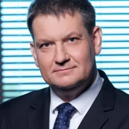 Portret biznesowy mężczyzny w ciemnym garniturze i wzorzystym krawacie na tle niebieskich żaluzji.