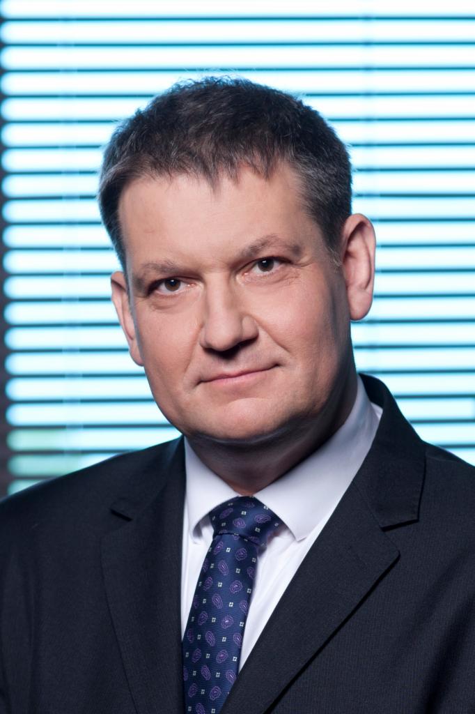 Portret biznesowy mężczyzny w ciemnym garniturze i wzorzystym krawacie na tle niebieskich żaluzji.