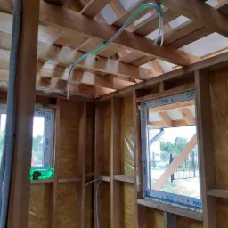 Wnętrze szkieletu drewnianego domu w trakcie budowy, widoczne kable elektryczne prowadzone pod sufitem oraz okna z zamontowaną stolarką okienną, poziomica laserowa ustawiona na parapecie.