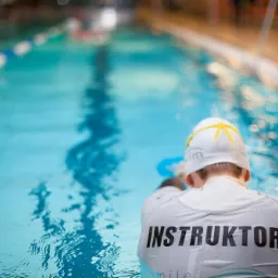 Instruktor pływania w białym stroju i czepku z żółtymi detalami stoi w basenie, obserwując pływających w oddali.