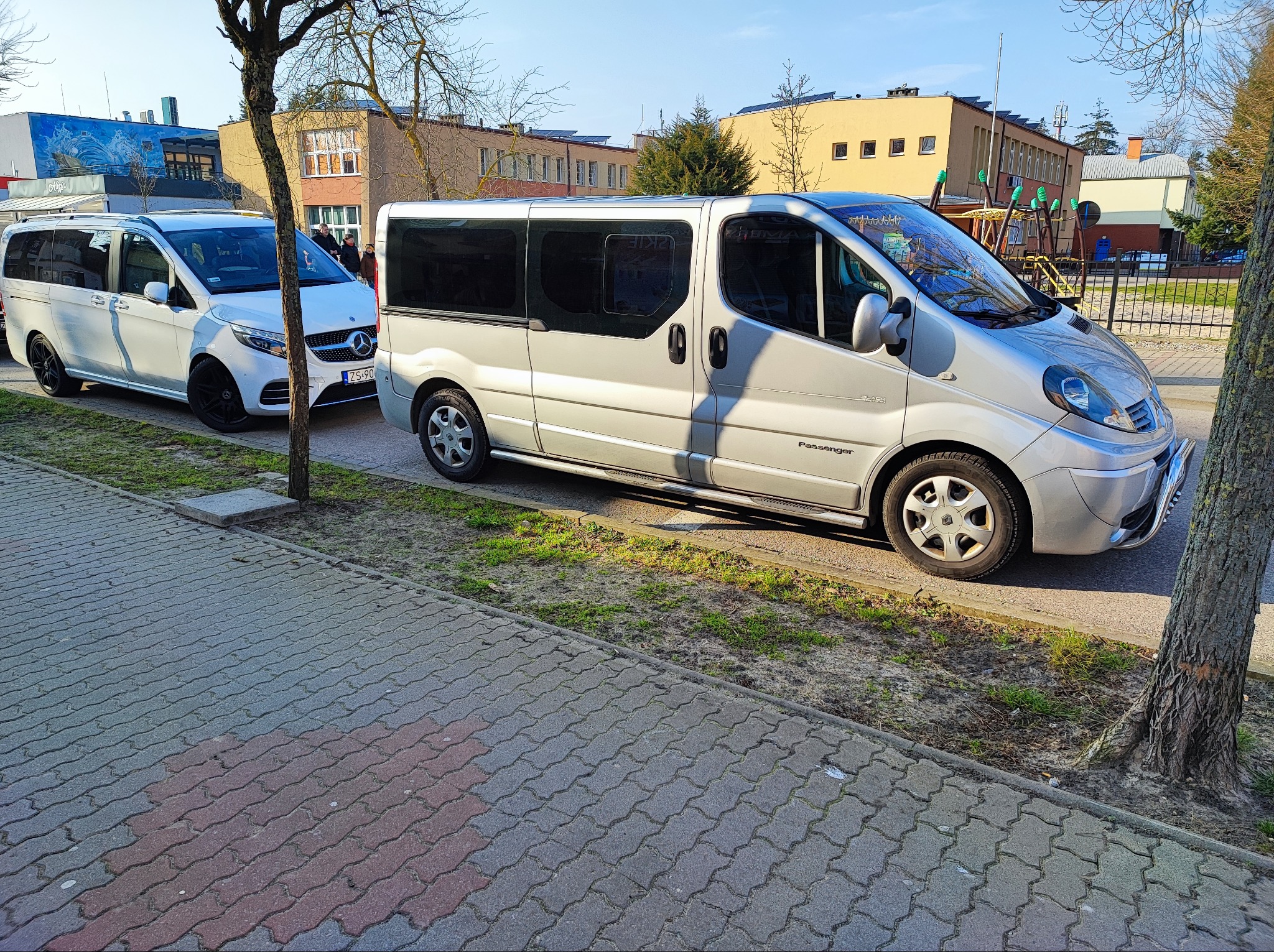 Dwa busy: biały Mercedes i srebrny Renault, zaparkowane obok chodnika z kostki brukowej i drzewa na tle budynków.