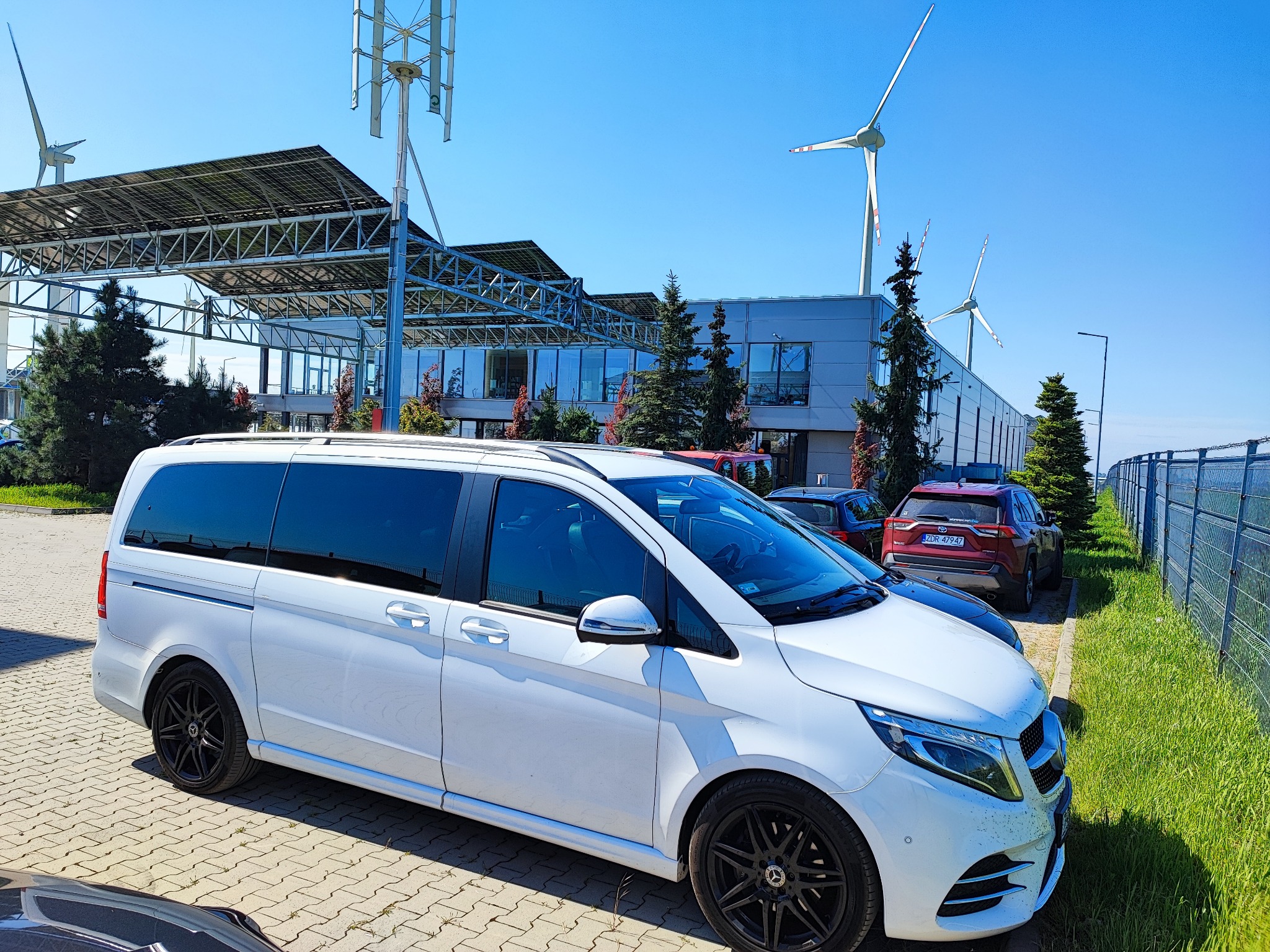 Biały van Mercedes V-klasy zaparkowany na brukowanym parkingu, z turbinami wiatrowymi i panelami słonecznymi w tle.