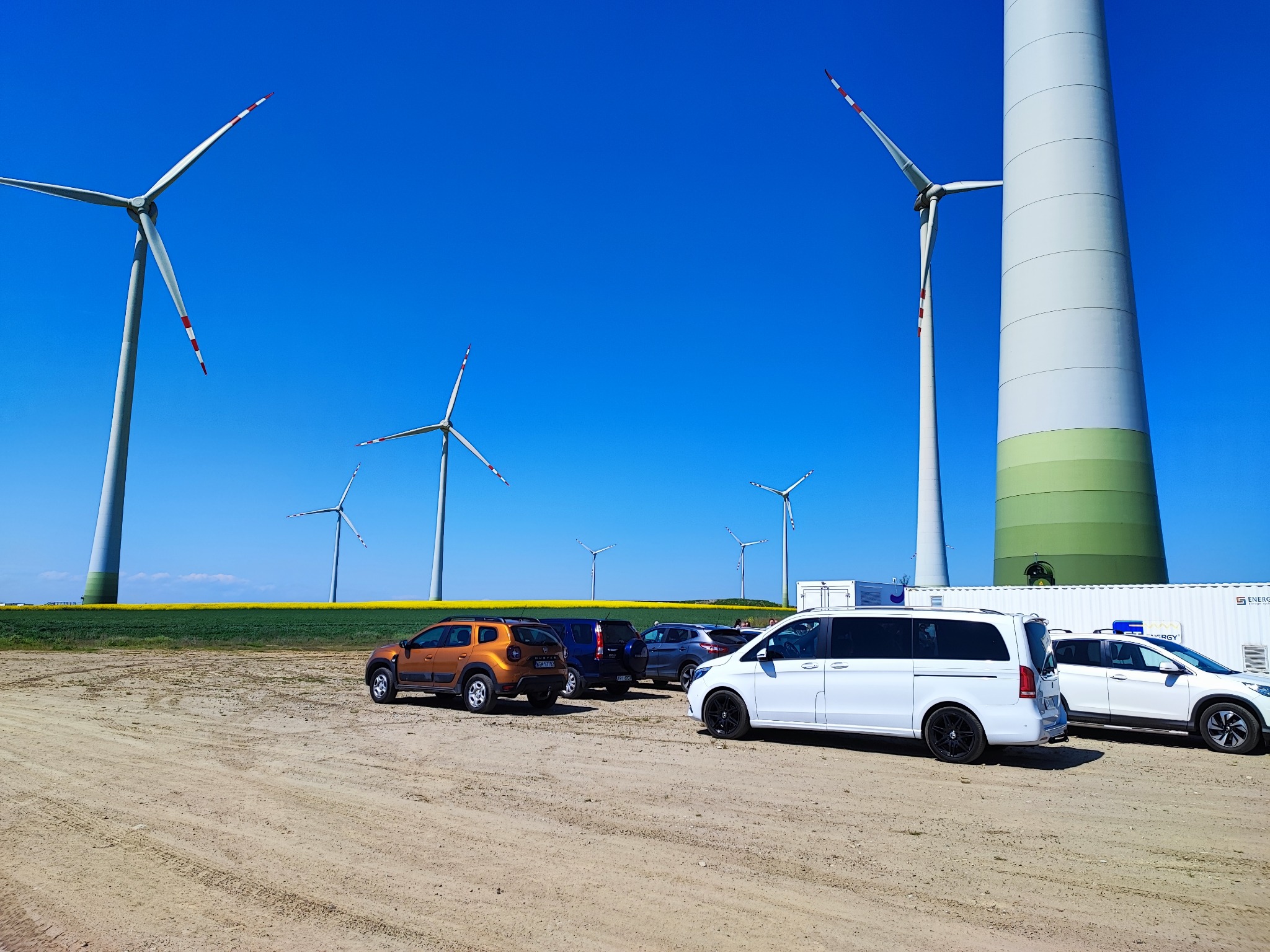 Parking z różnymi samochodami osobowymi, w tle turbiny wiatrowe i pole rzepaku.
