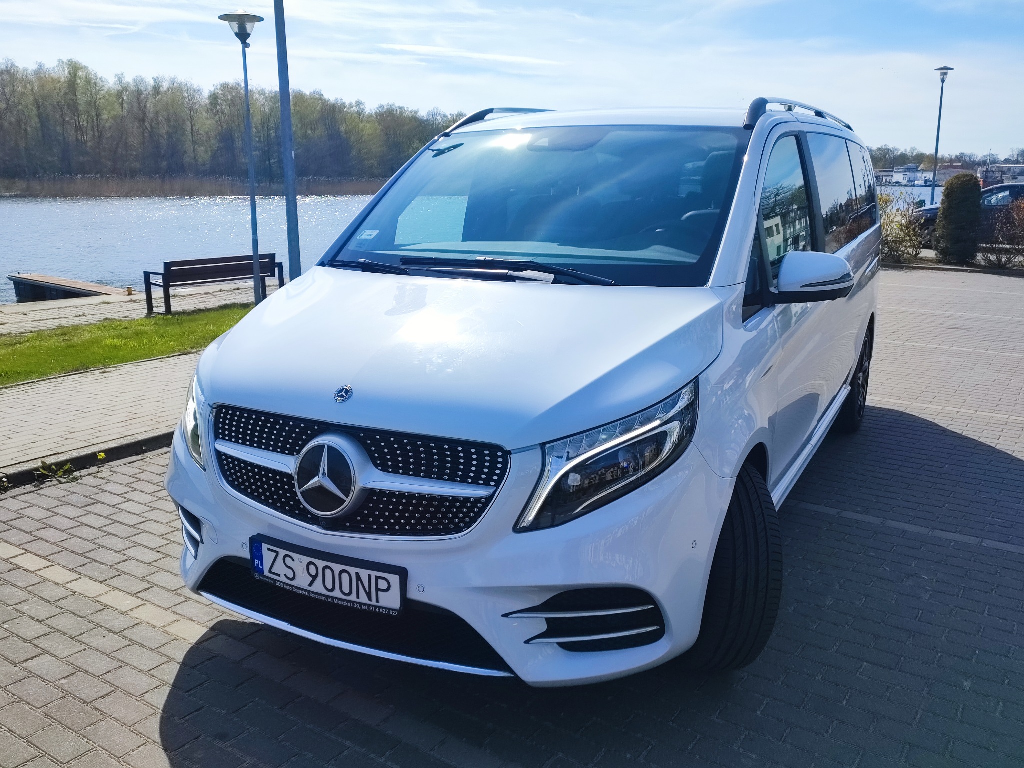 Biały Mercedes V-klasy zaparkowany na brukowanym placu z widokiem na rzekę i drzewa w tle, tablica rejestracyjna ZS 900NP.