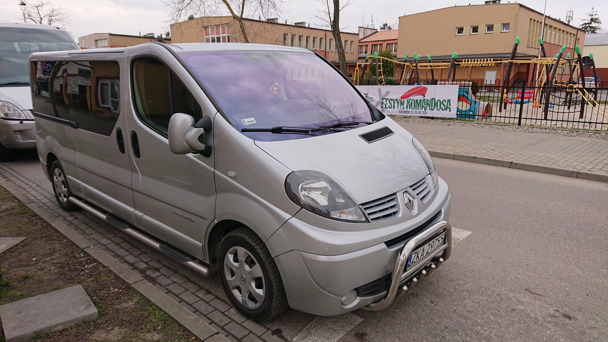 Srebrny van Renault Trafic z relingami i chromowanym orurowaniem parkuje na ulicy, w tle plac zabaw i budynek szkolny.