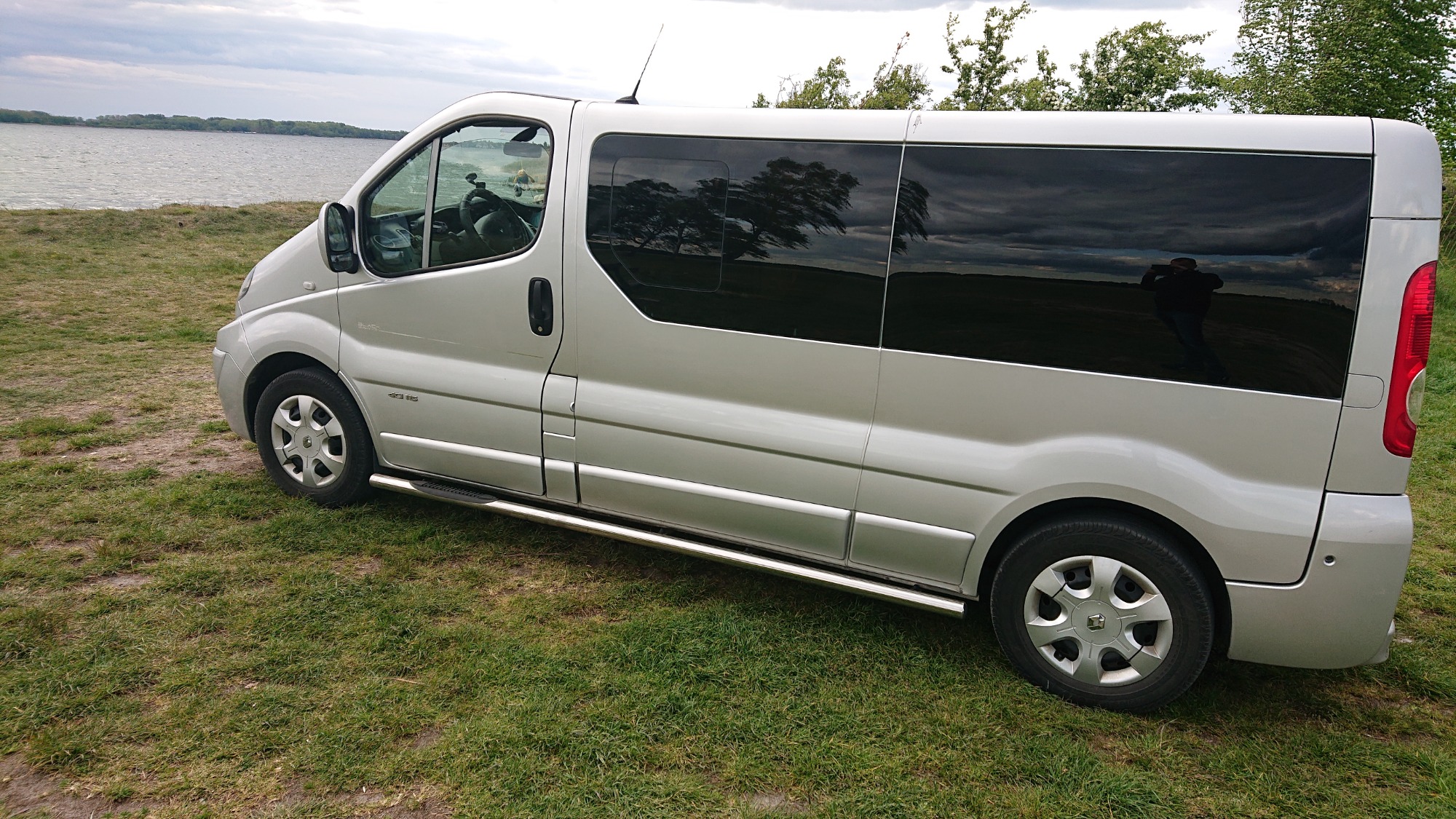 Srebrny bus z przyciemnianymi szybami zaparkowany na trawie, z widocznym odbiciem osoby w oknie i jeziorem w tle.
