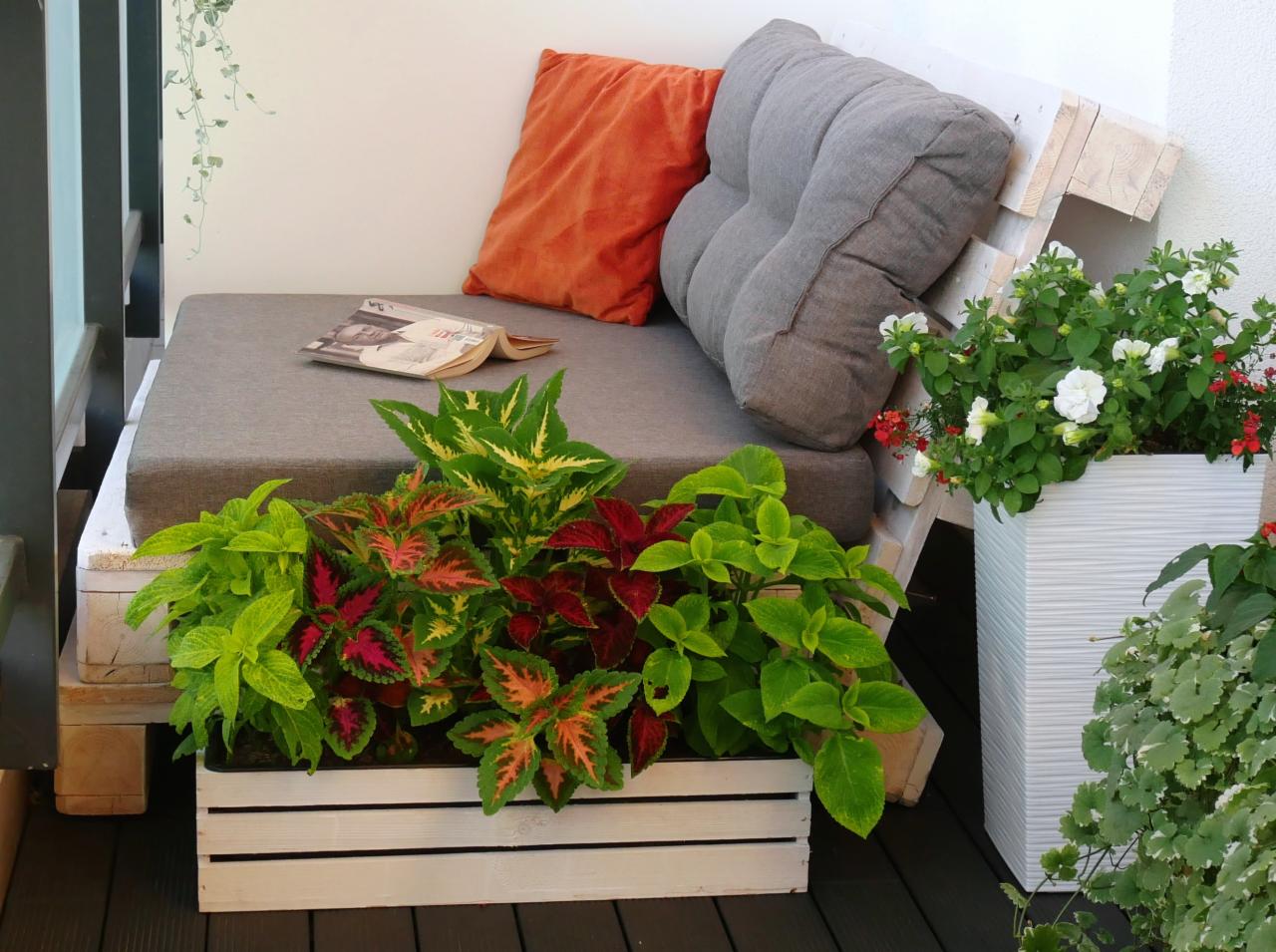 Aranżacja balkonu z siedziskiem z palet, poduszką w kolorze pomarańczowym, szarym podparciem pleców i roślinami w skrzynkach i donicach.