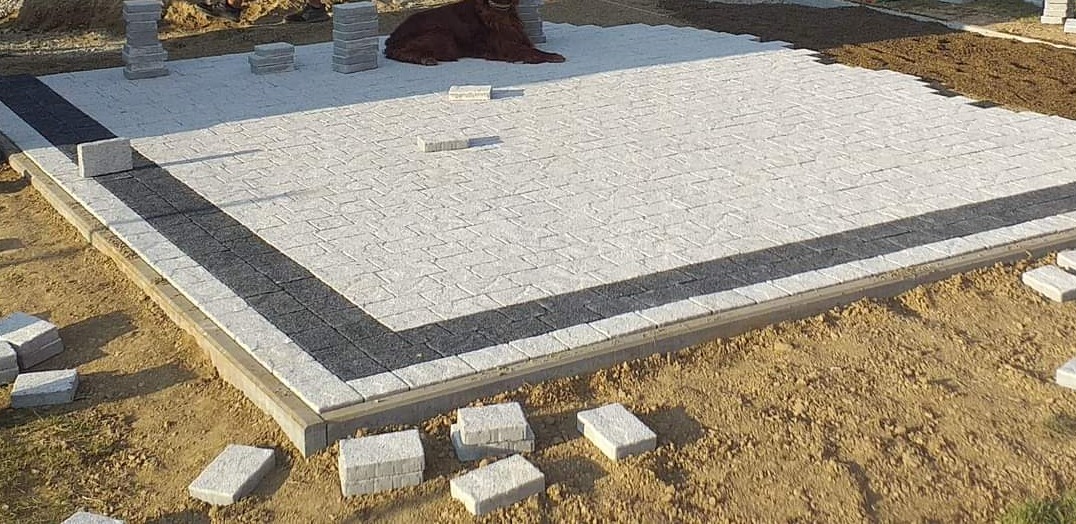 Fragment powierzchni z ułożoną jasną kostką brukową w geometryczny wzór, obramowaną ciemniejszym pasem, z rudym psem leżącym na środku, w trakcie prac wykończeniowych.