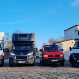 Cztery busy: dwa białe Renault Master z zabudową kontenerową, jeden ciemny Peugeot Boxer z zabudową Lamar i czerwony Volkswagen Transporter T4, ustawione obok siebie na placu z kostki brukowej...