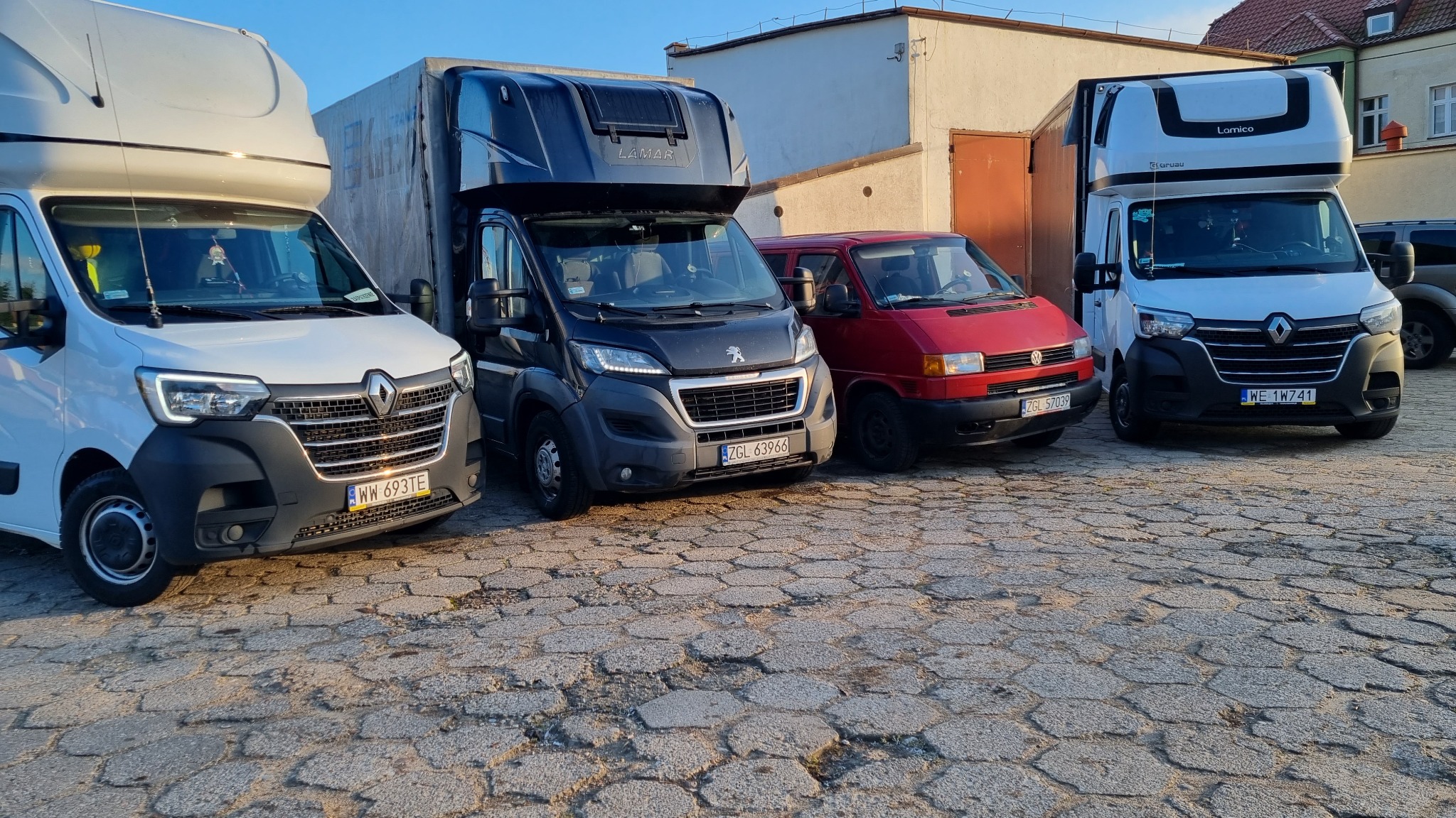 Flota różnych busów dostawczych z wysoką zabudową zaparkowana na brukowanym placu, w tle budynek z pomarańczowymi drzwiami garażowymi.