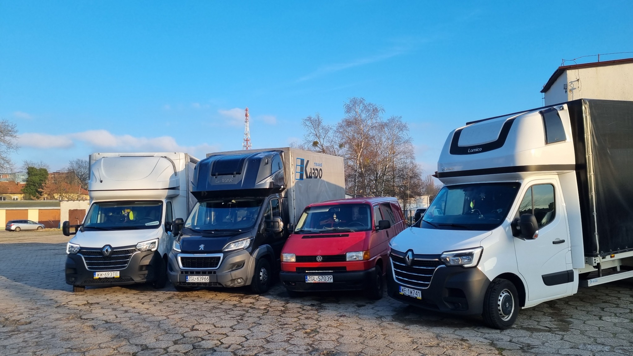 Cztery busy transportowe różnych marek (Renault, Peugeot, Volkswagen) zaparkowane obok siebie na brukowanym placu w słoneczny dzień. Widoczne tablice rejestracyjne i zabudowy kontenerowe.