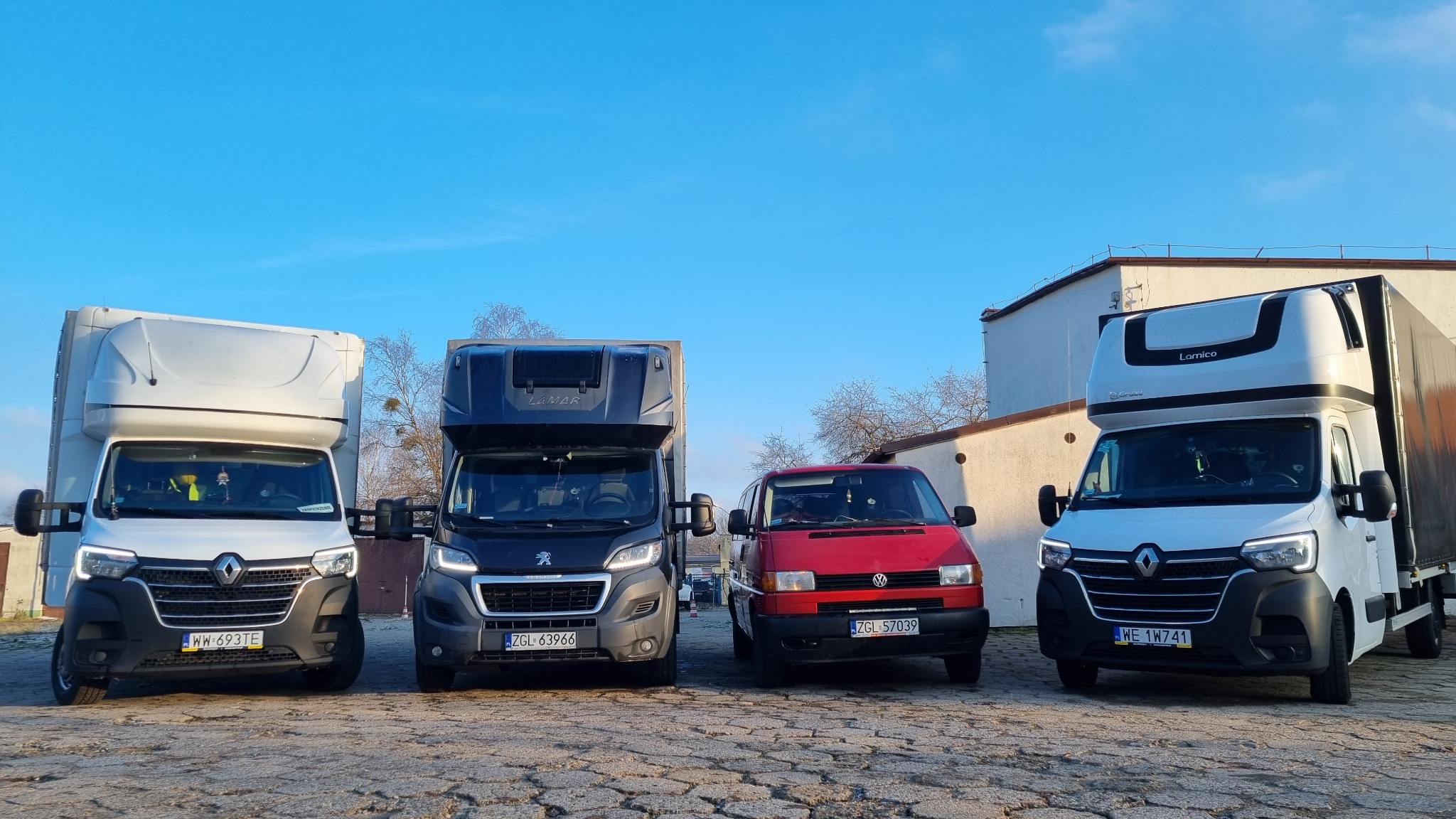Cztery busy: dwa białe Renault Master z zabudową kontenerową, jeden ciemny Peugeot Boxer z zabudową Lamar i czerwony Volkswagen Transporter T4, ustawione obok siebie na placu z kostki brukowej...