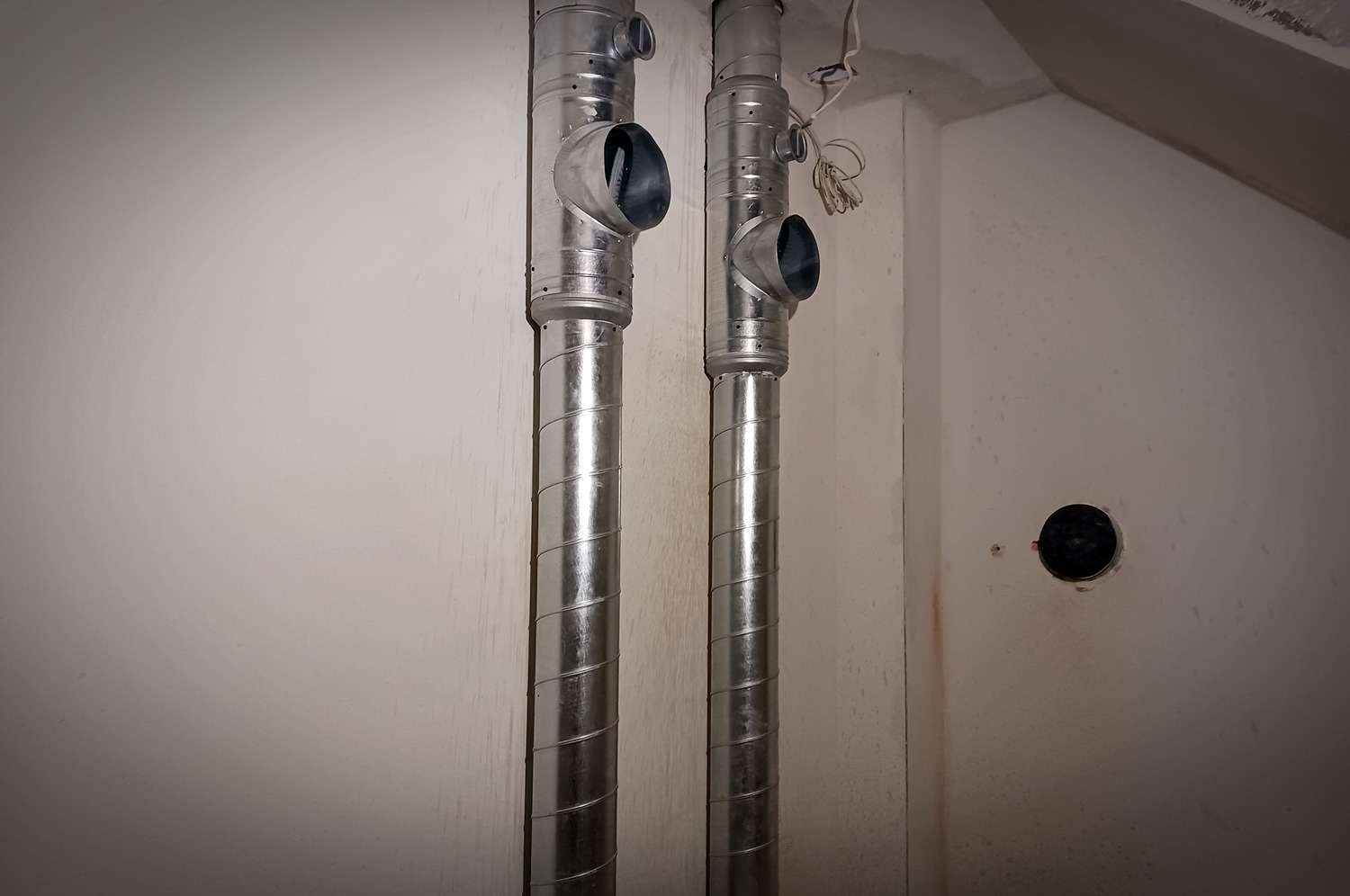Dwie pionowe, spiralnie zwijane rury wentylacyjne z odgałęzieniami, zamontowane przy białej ścianie w surowym stanie, widoczne przewody i otwór w ścianie.