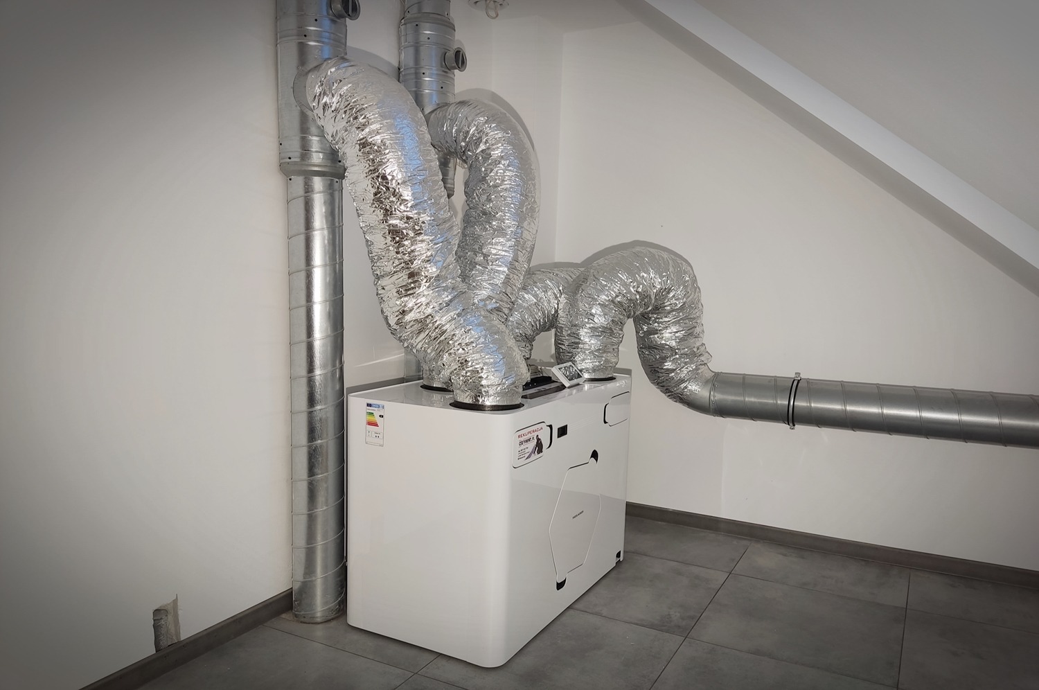 Biała jednostka centralna z podłączonymi, srebrnymi, elastycznymi przewodami wentylacyjnymi w pomieszczeniu z szarą podłogą z płytek.
