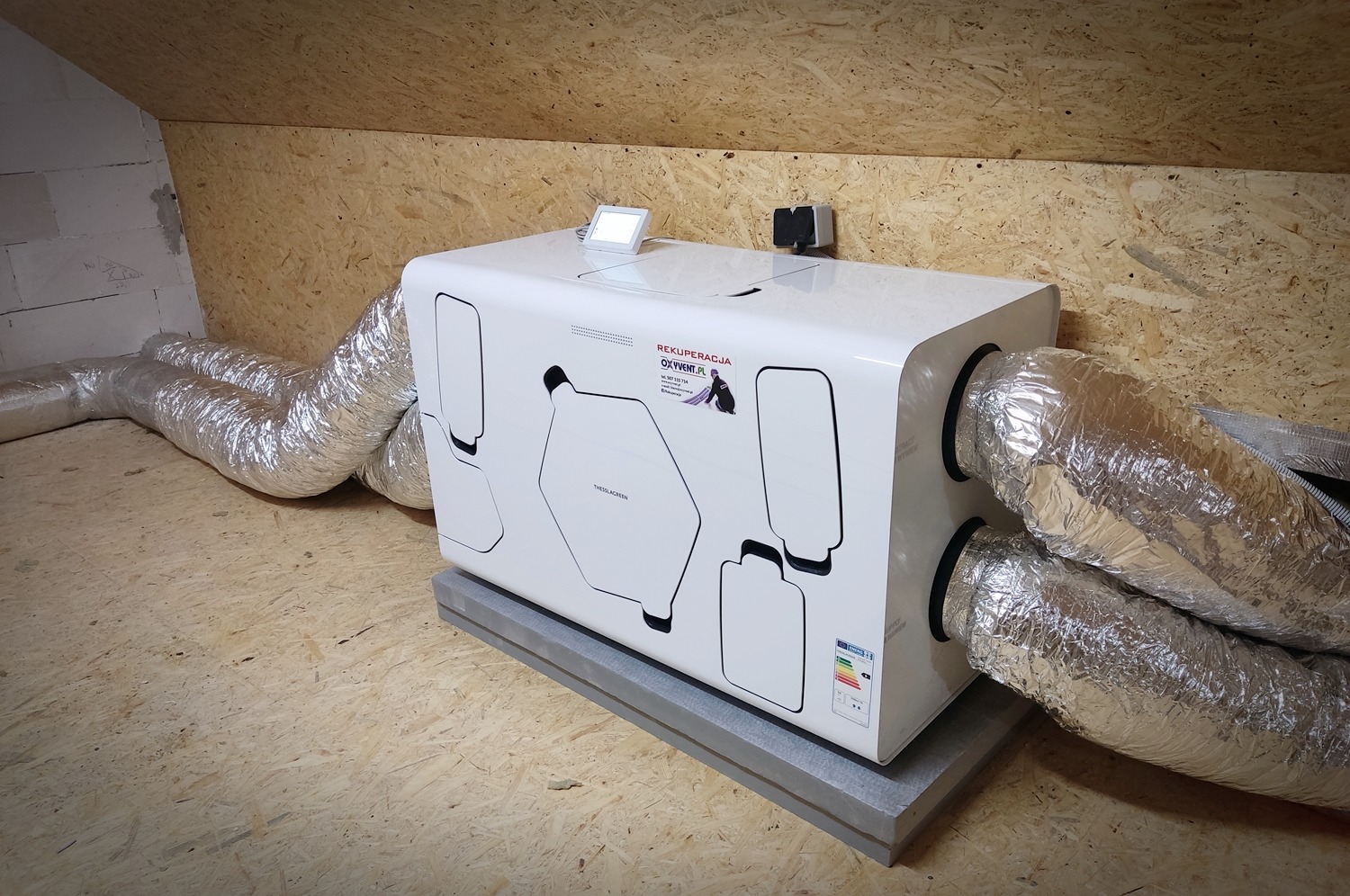 Biała centrala wentylacyjna z podłączonymi przewodami wentylacyjnymi w pomieszczeniu na poddaszu, widoczne elementy sterujące i naklejki informacyjne.