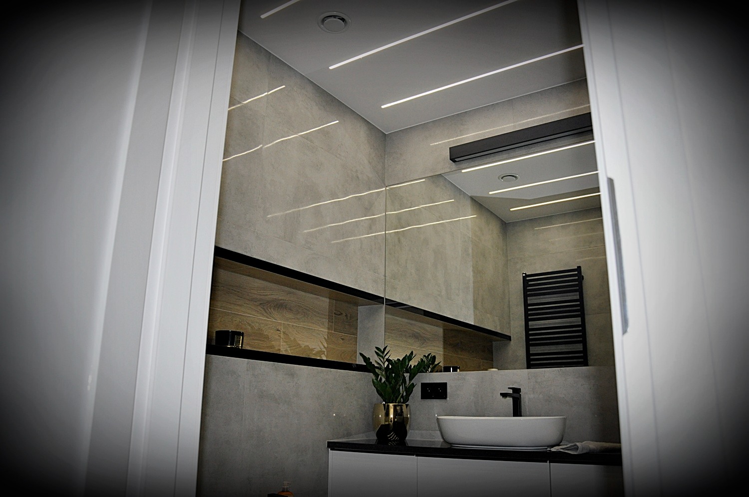 Nowoczesna łazienka z oświetleniem liniowym LED w Parczewie, widoczna przez otwarte drzwi, z umywalką nablatową, grzejnikiem drabinkowym i półką z drewnianym akcentem.