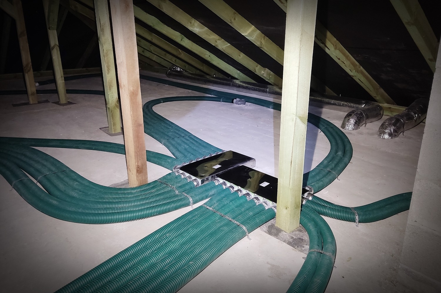 Montaż systemu rekuperacji z zielonymi przewodami wentylacyjnymi w przestrzeni poddasza, widoczne skrzynki rozdzielcze i drewniana konstrukcja dachu.