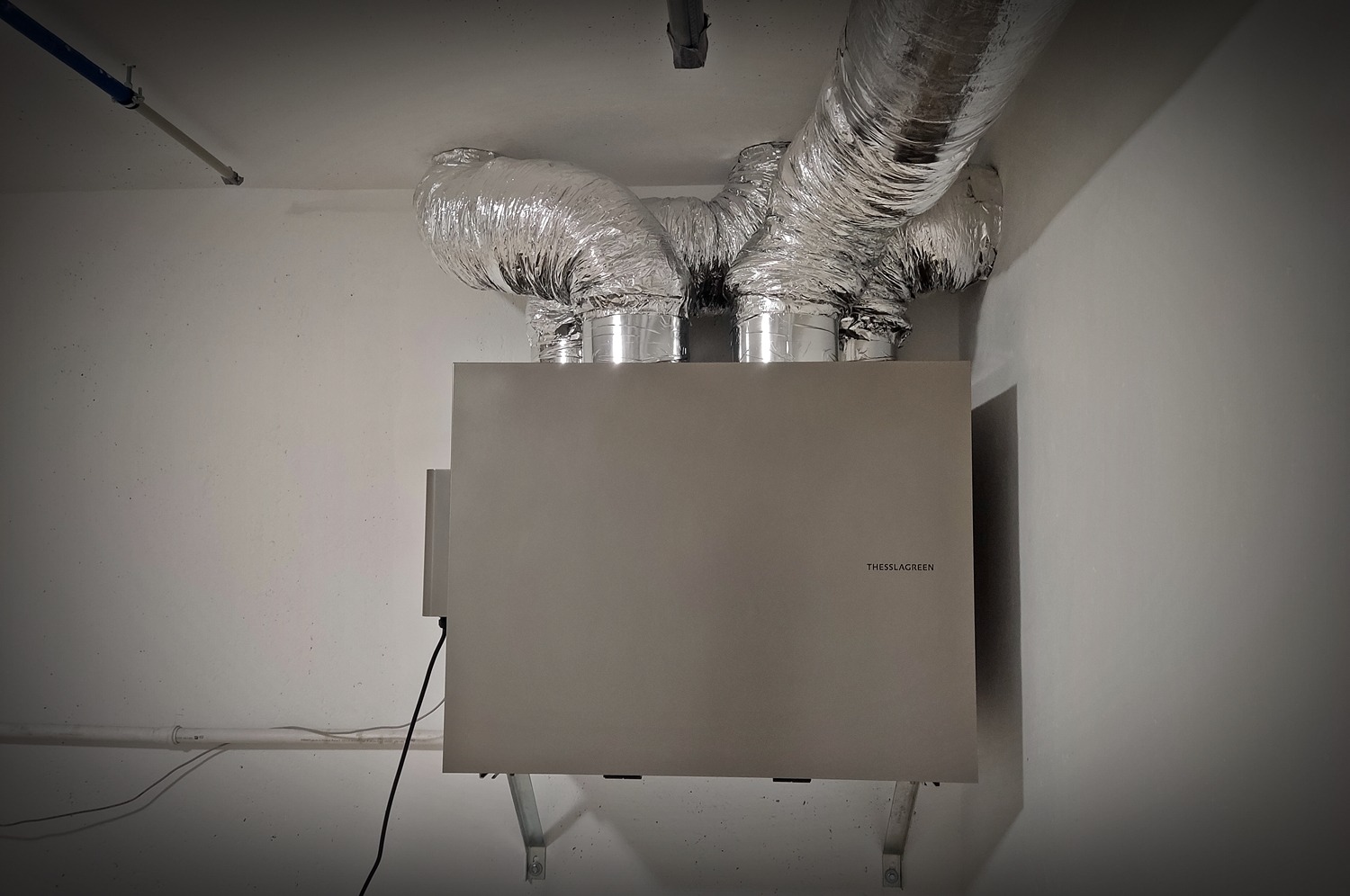 Szara centrala wentylacyjna z podłączonymi, elastycznymi przewodami wentylacyjnymi, zamontowana na ścianie pomieszczenia technicznego z widocznymi rurami instalacyjnymi i napisem THESSLAGREEN.