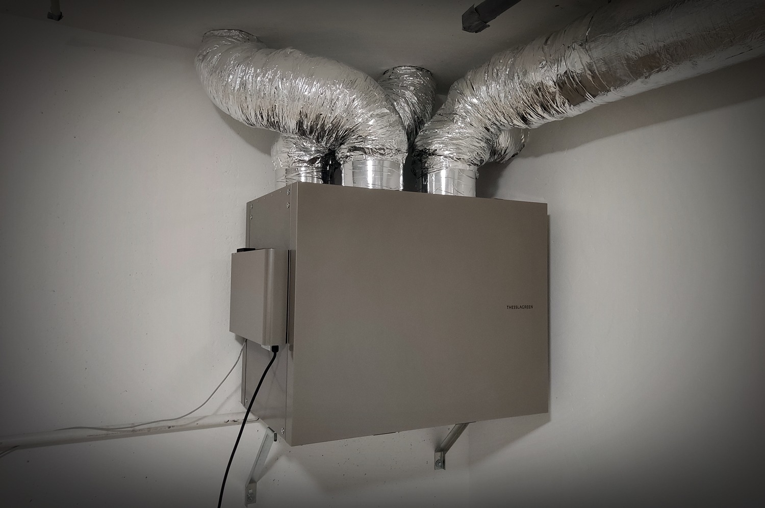 Szara centrala wentylacyjna z trzema elastycznymi, aluminiowymi przewodami wentylacyjnymi podłączonymi u góry, zamontowana na ścianie z widocznymi przewodami elektrycznymi.