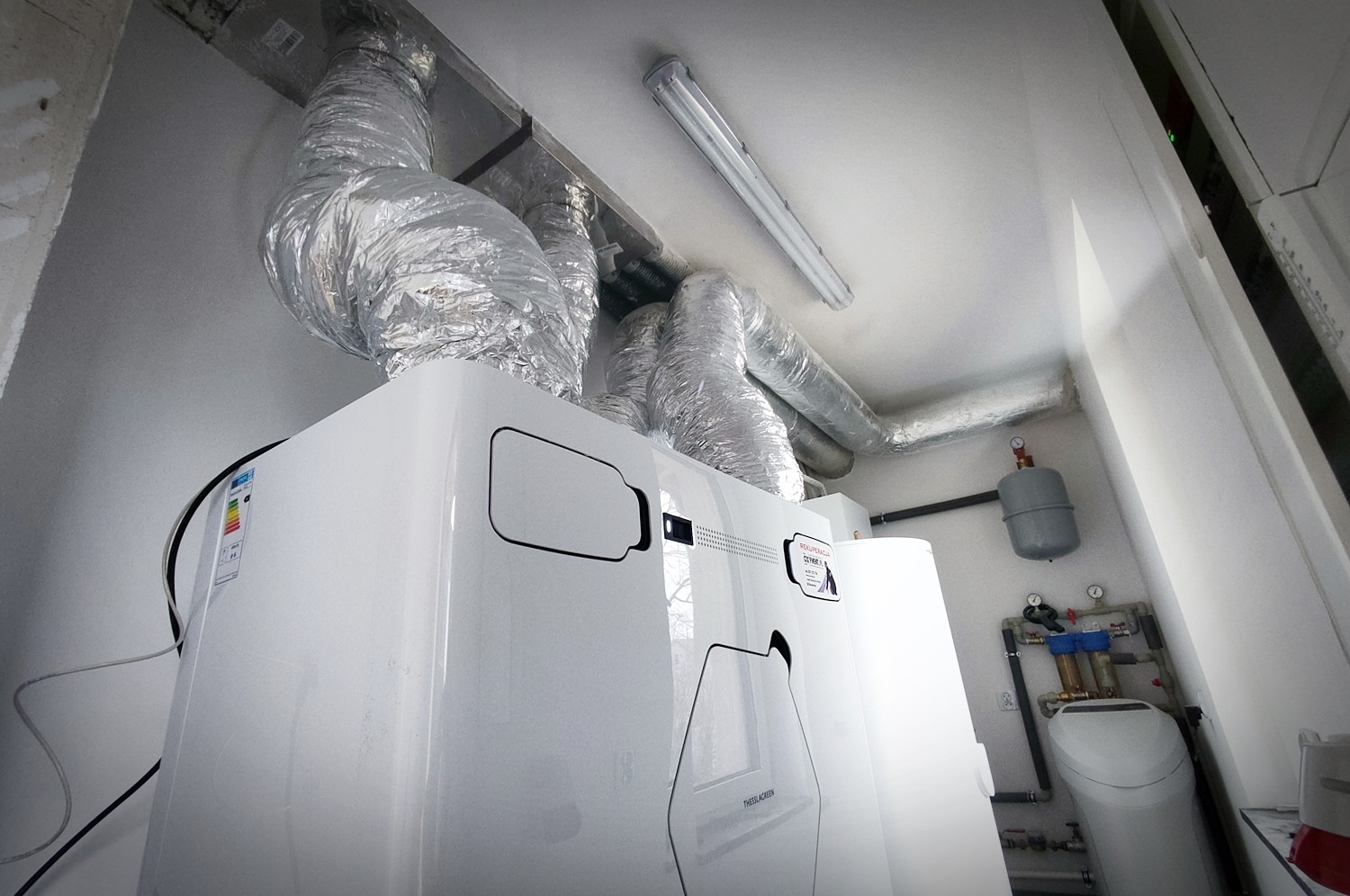 Biała jednostka centralna z podłączonymi izolowanymi przewodami wentylacyjnymi, widoczne elementy instalacji hydraulicznej na ścianie, w tle oświetlenie sufitowe.