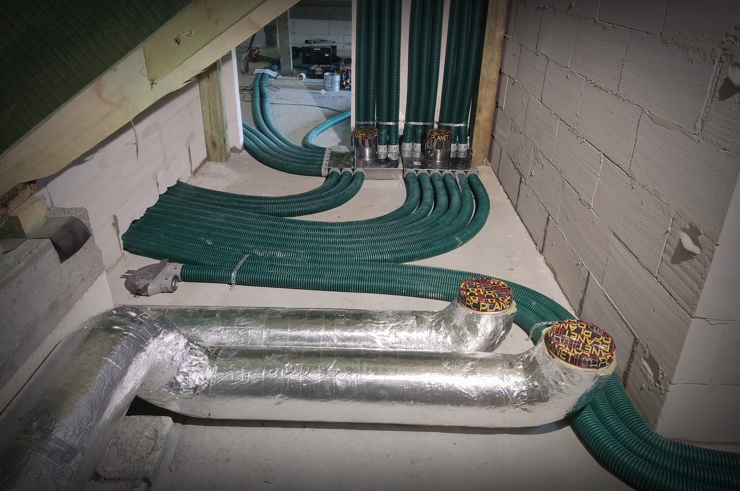 Wnętrze poddasza z widocznymi zielonymi rurami wentylacyjnymi, niektóre owinięte srebrną folią izolacyjną, prowadzącymi do metalowych rozdzielaczy. Rury układają się w równoległe linie,...