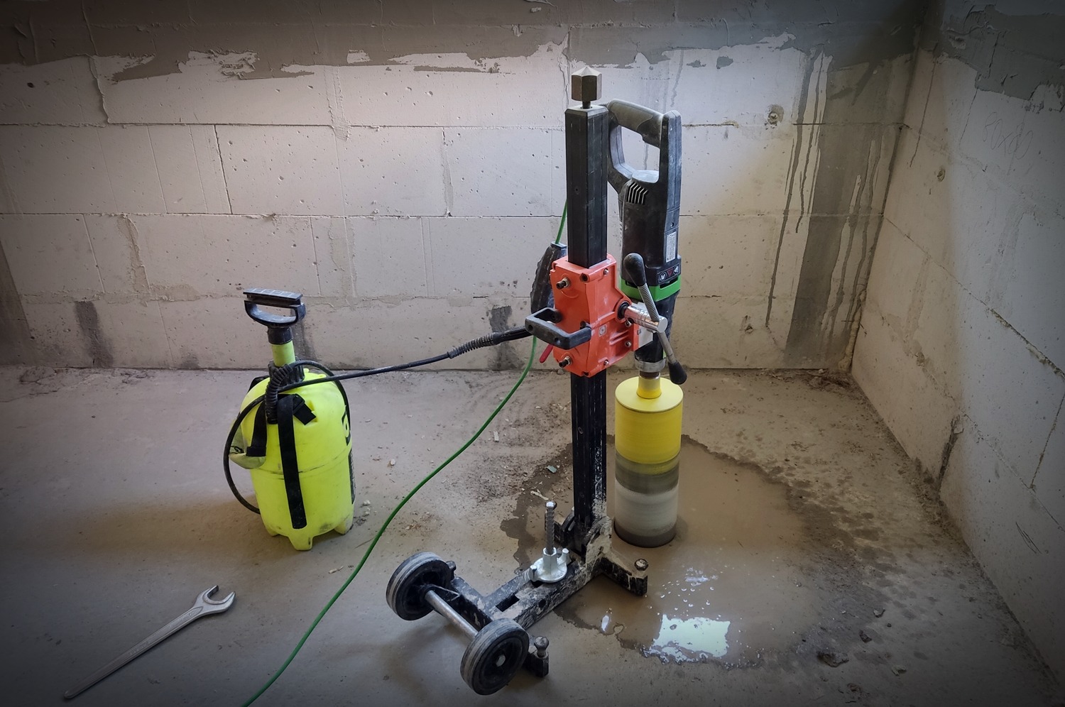 Wiertnica diamentowa na mokro z żółtą koronką, wiertło w trakcie wiercenia otworu w posadzce w surowym pomieszczeniu z bloczków betonowych, obok spryskiwacz ciśnieniowy i klucz płaski.