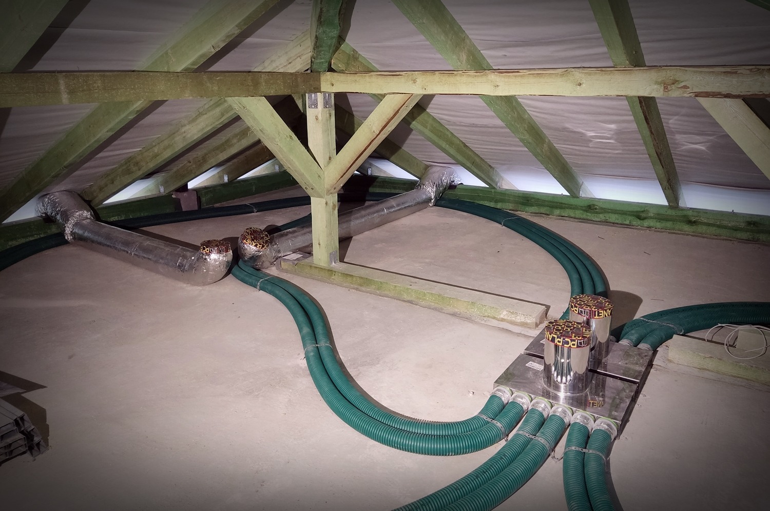 Instalacja systemu wentylacyjnego z rurami w kolorze zielonym i srebrnym, widoczne poddasze z drewnianą konstrukcją, centrala wentylacyjna z czterema przyłączami.