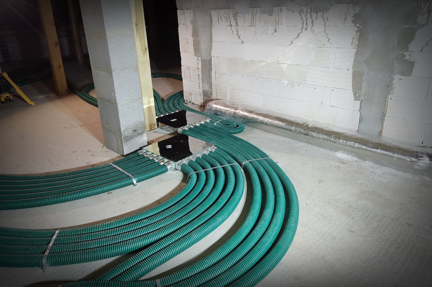 Rozprowadzenie systemu rekuperacji w surowym stanie budynku, widoczne zielone rury wentylacyjne, srebrna rura izolowana, betonowe ściany i filary w trakcie budowy.