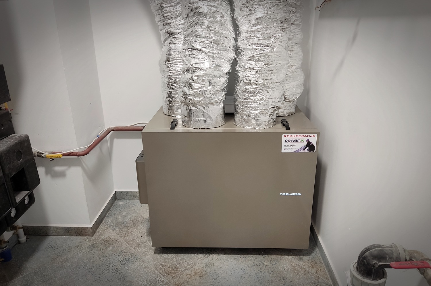 Centrala wentylacyjna z czterema przewodami wentylacyjnymi w srebrnej izolacji, widoczna etykieta z danymi firmy i logo, oraz fragment instalacji wodnej w tle.