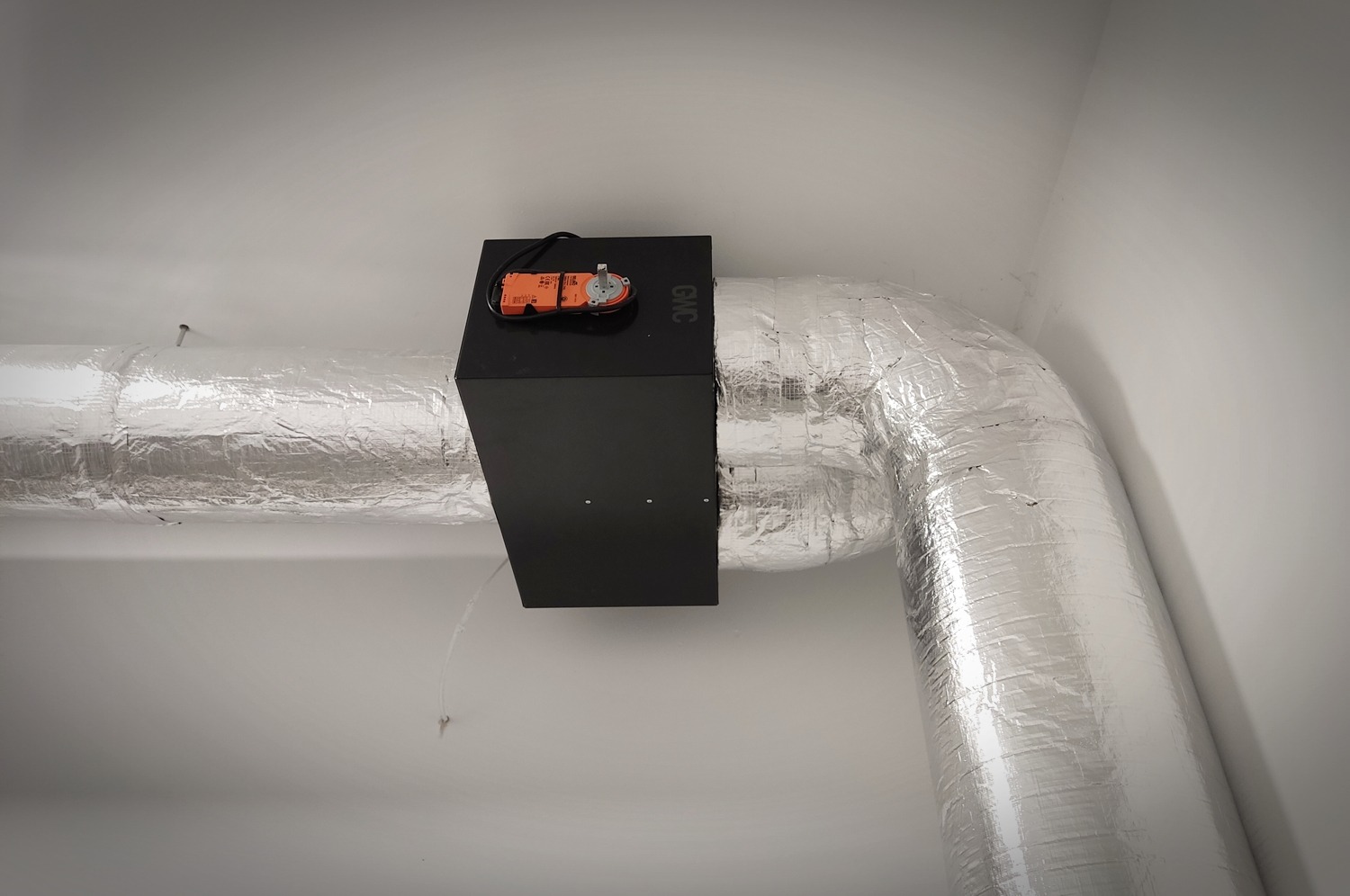 Fragment instalacji wentylacyjnej z izolowanym kanałem, czarną skrzynką z napędem i widocznym przewodem, zamontowanej pod sufitem w rogu pomieszczenia.