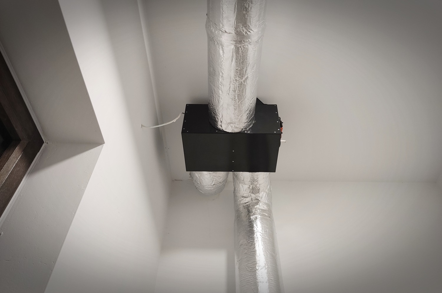 Czarna centrala wentylacyjna z aluminiowymi przewodami wentylacyjnymi zamontowana pod sufitem w rogu jasnego pomieszczenia z widocznym fragmentem okna.