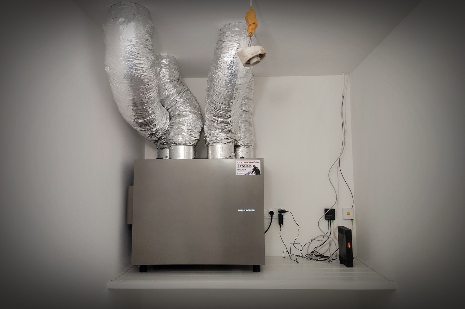 Szara jednostka centralna z podłączonymi, srebrnymi, izolowanymi przewodami wentylacyjnymi w białym pomieszczeniu, widoczny panel sterowania i naklejka firmy.