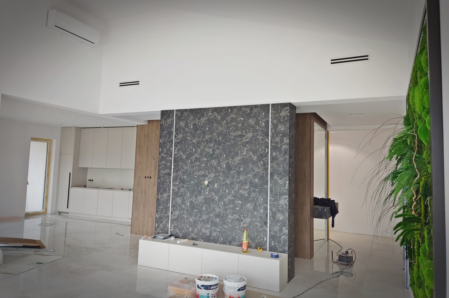 Wnętrze salonu w trakcie wykańczania z widoczną instalacją wentylacyjną w suficie, marmurową ścianą, panelami z drewna, ścianą z mchu i elementami instalacji elektrycznej na podłodze.