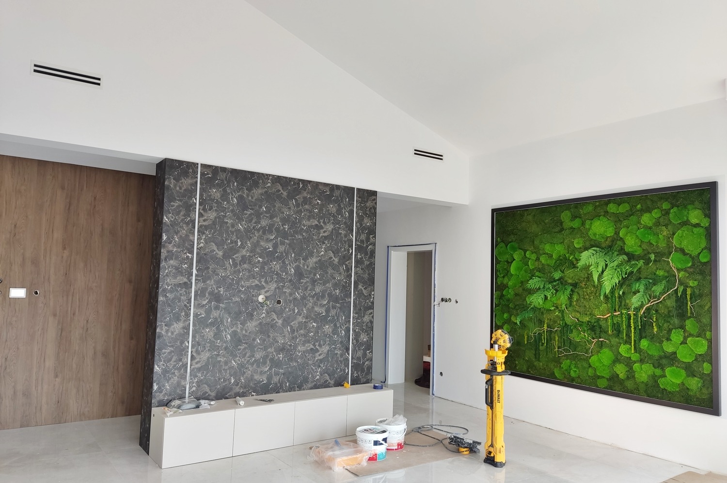 Wnętrze w trakcie prac wykończeniowych: widoczne kanały wentylacyjne w suficie, ściana z okładziną imitującą marmur, szafka RTV, obraz z mchu na ścianie oraz narzędzia pomiarowe i puszki z farbą...
