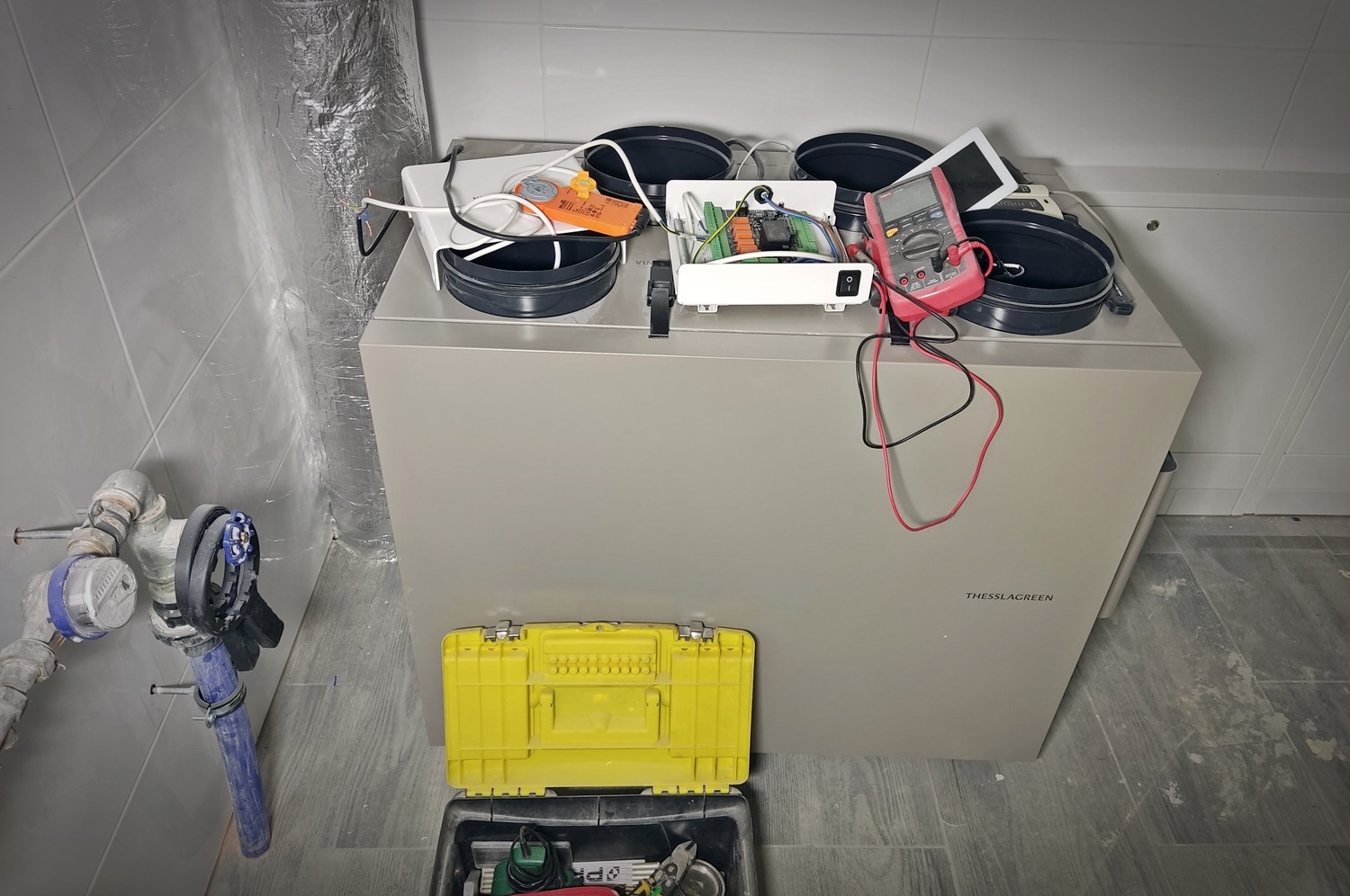 Szary rekuperator Thesslagreen z otwartą obudową elektroniki, multimetrem, czarnymi okrągłymi elementami, żółtą skrzynką narzędziową i rurami wodnymi w tle.