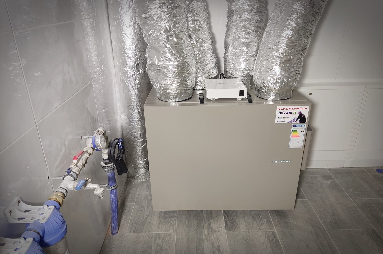 Szara centrala wentylacyjna z podłączonymi elastycznymi przewodami aluminiowymi, widoczne przyłącza wodne z filtrem i zaworami, naklejka informacyjna na obudowie, płytki ceramiczne na ścianach...