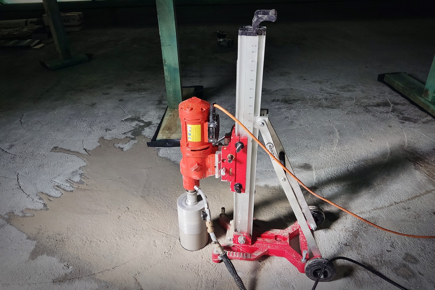 Czerwona wiertnica rdzeniowa z mokrym wierceniem w betonie, widoczna woda po wierceniu, podpora pionowa z miarką, kabel zasilający, w tle fragmenty drewnianej konstrukcji.