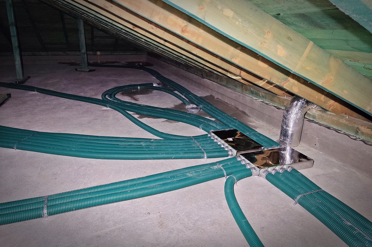 Zielone, karbowane rury wentylacyjne rozprowadzone w przestrzeni poddasza, podłączone do metalowych rozdzielaczy i izolowanego przewodu pionowego.