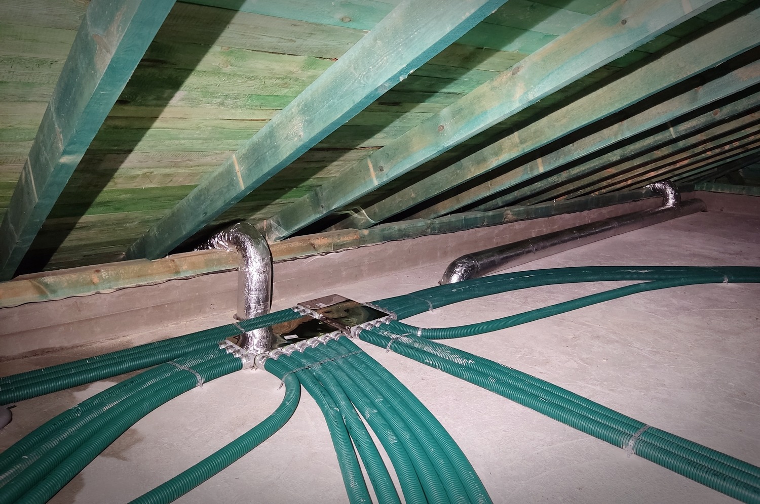 Rozprowadzenie zielonych, karbowanych przewodów wentylacyjnych z metalowej skrzynki rozdzielczej na poddaszu, widoczne srebrne rury wentylacyjne pod skośnym, zielonym dachem z desek.