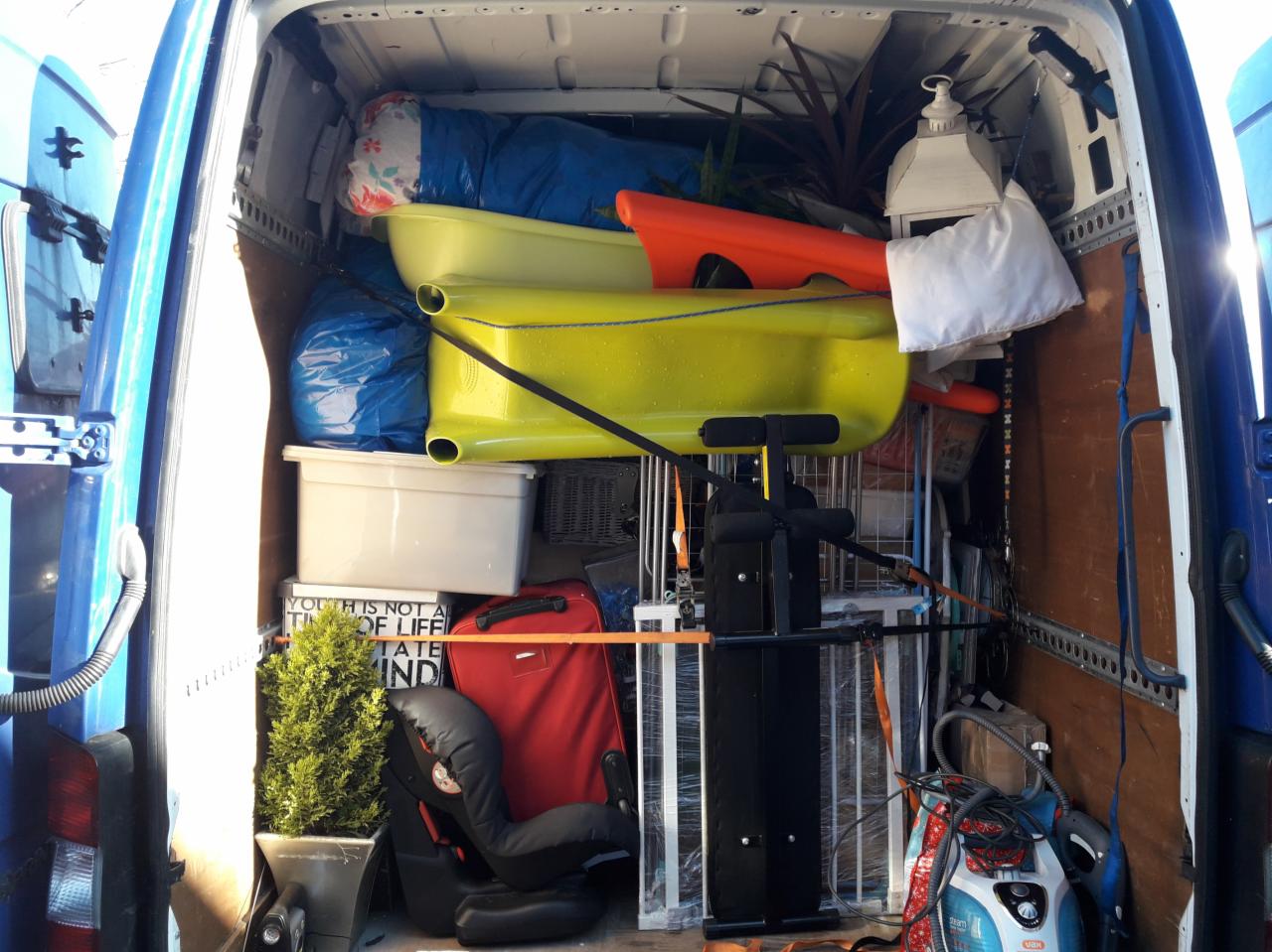 Wnętrze furgonetki załadowane różnorodnymi przedmiotami podczas przeprowadzki, w tym wanienki dziecięce, walizka, fotelik samochodowy i sprzęt do ćwiczeń, zabezpieczone pasami.