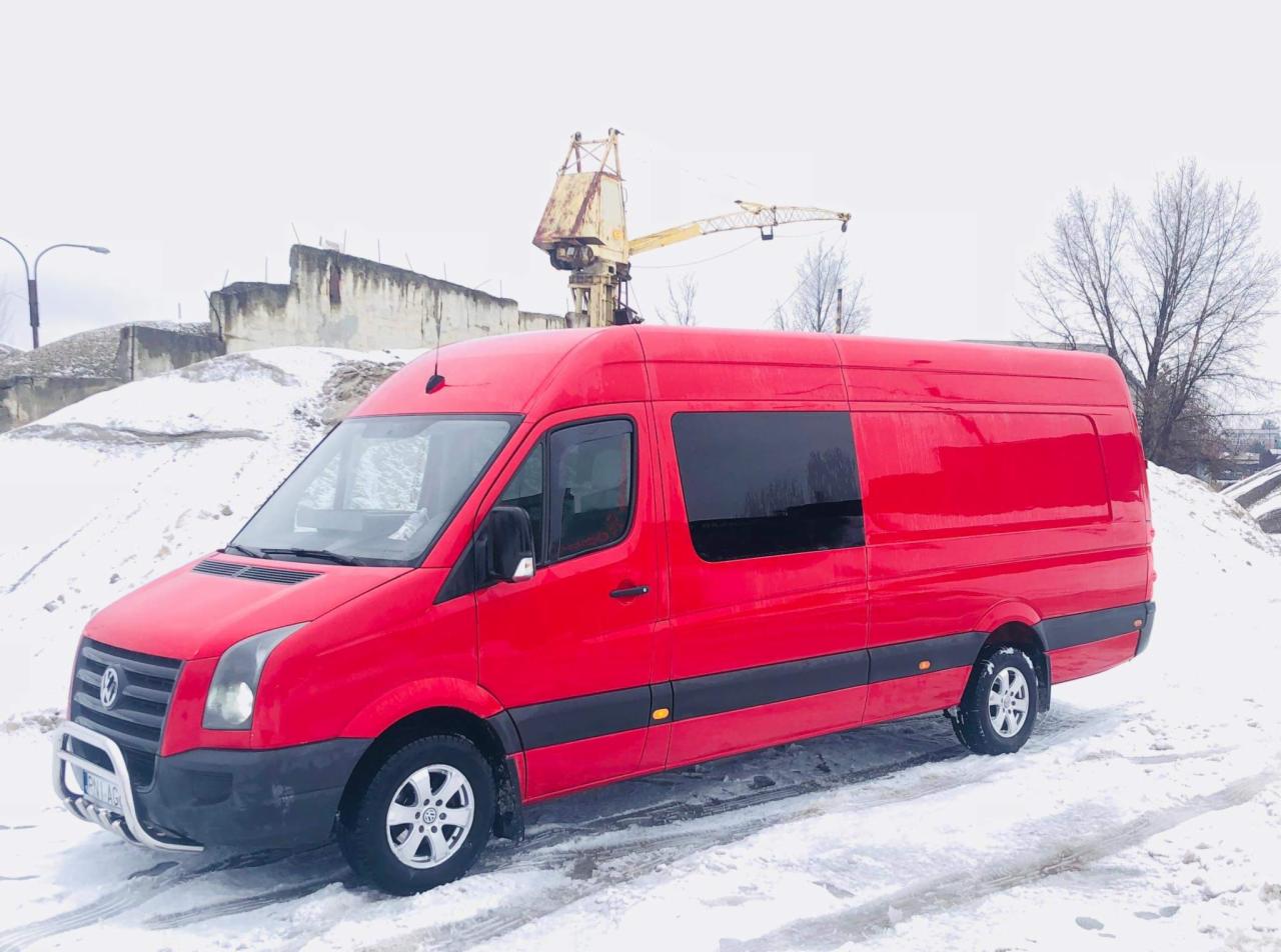 Czerwony bus transportowy na śniegu, z dźwigiem w tle i betonowymi konstrukcjami, widok z boku.