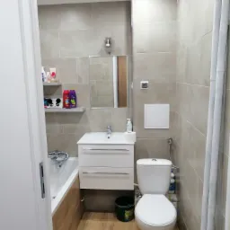 Wykończona łazienka z beżowymi płytkami, białą armaturą, wanną z obudową imitującą drewno i półkami z kosmetykami.