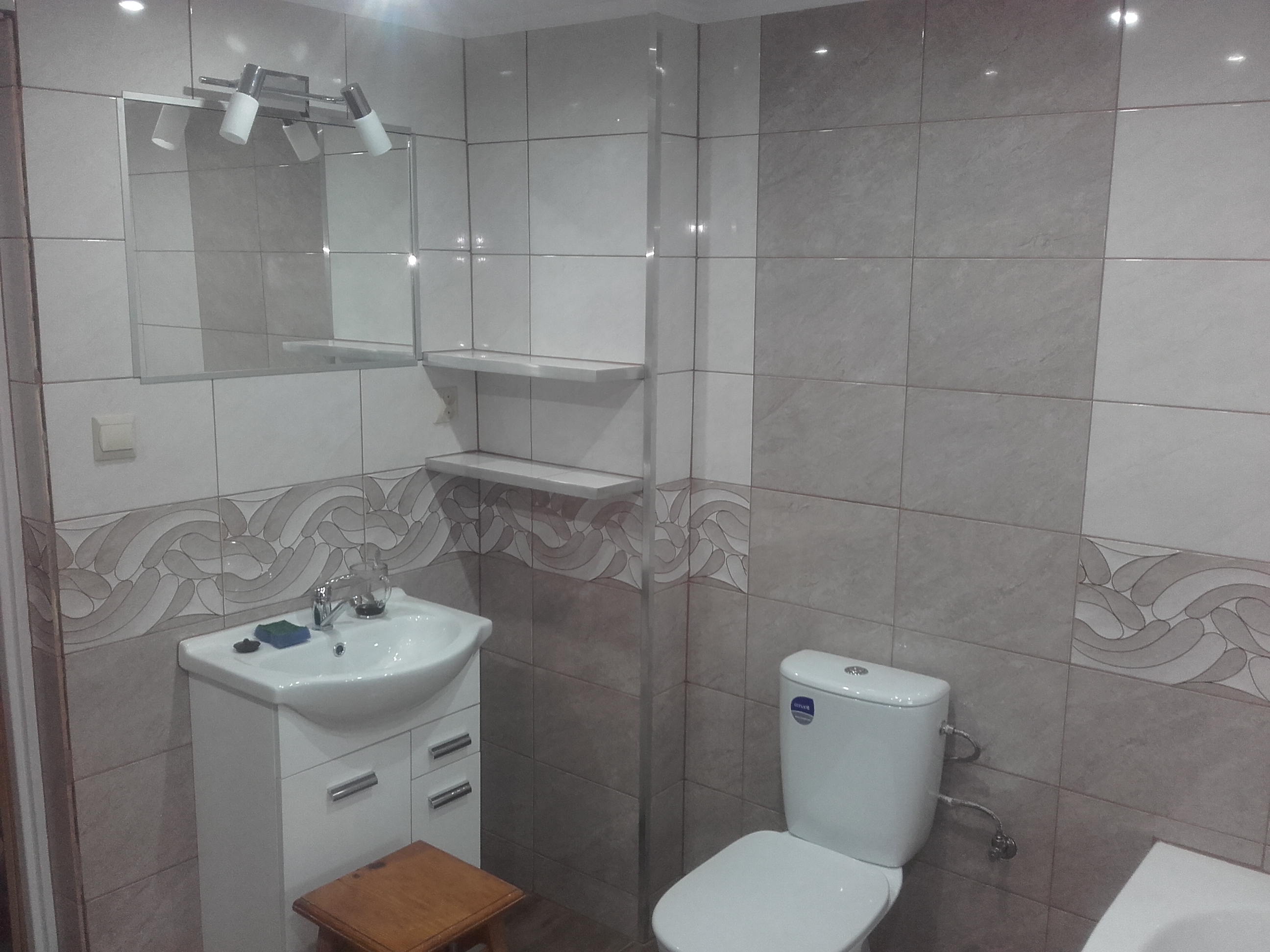 Wykończona łazienka z toaletą, umywalką z szafką, lustrem z oświetleniem i półkami, wyłożona płytkami w odcieniach szarości i bieli, z dekoracyjnym pasem płytek z motywem falistym.