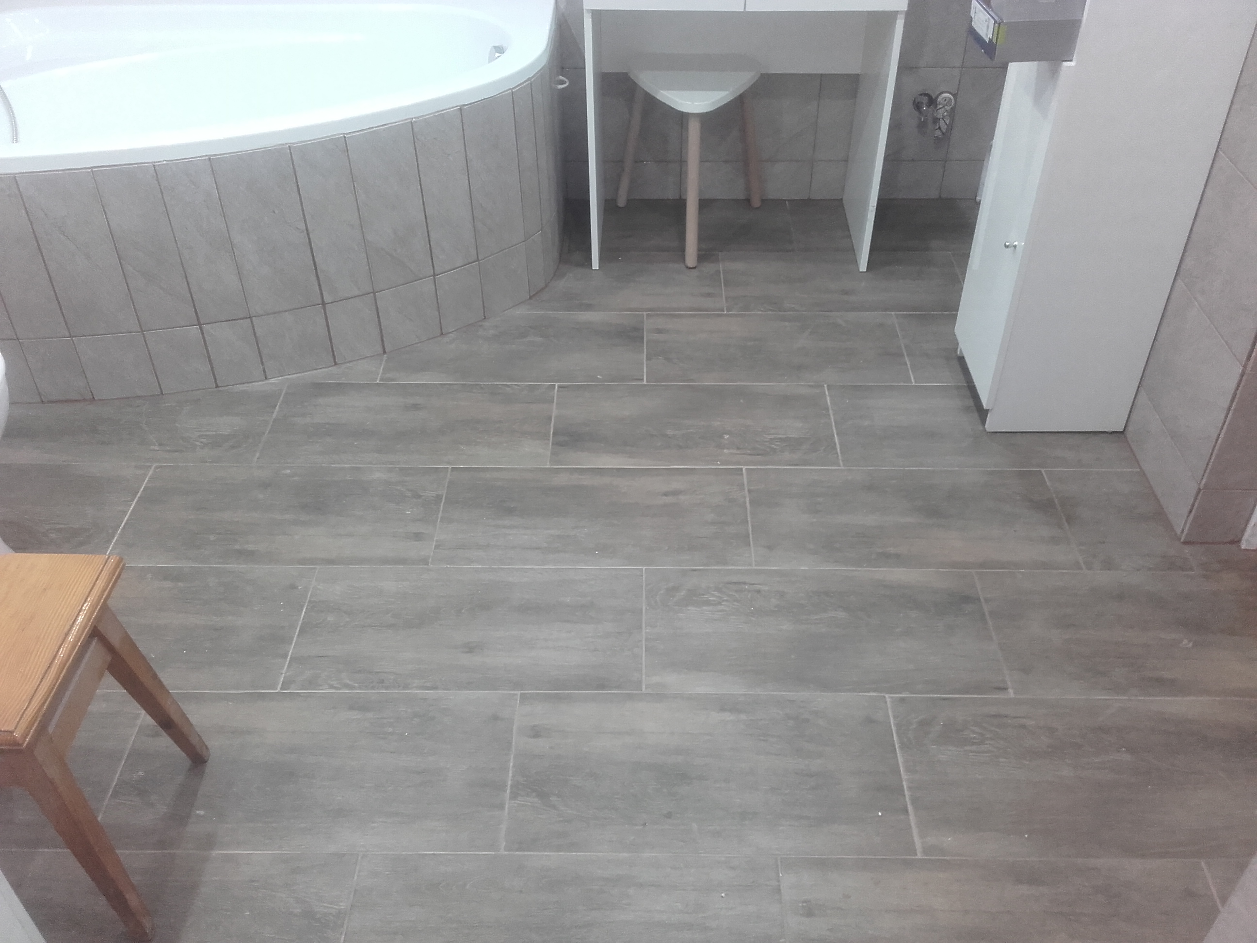 Ułożone szare płytki podłogowe imitujące drewno w łazience, widoczna wanna w zabudowie z płytek, biała szafka pod umywalką z krzesłem i fragment drewnianego taboretu.