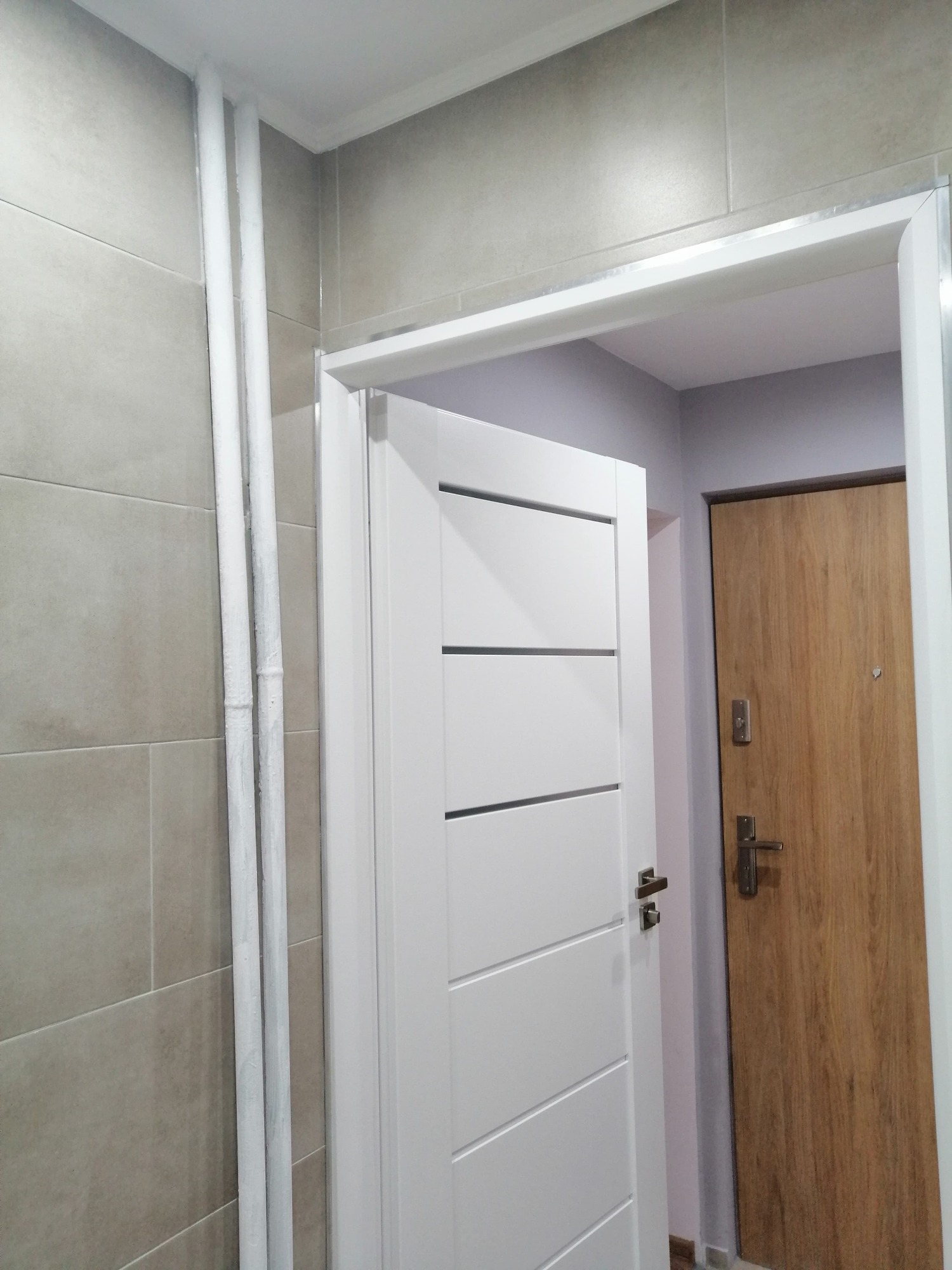 Wykończona łazienka z beżowymi płytkami, białą ościeżnicą i częściowo otwartymi białymi drzwiami z poziomymi frezami, widoczne rury pomalowane na biało oraz brązowe drzwi wejściowe w tle.